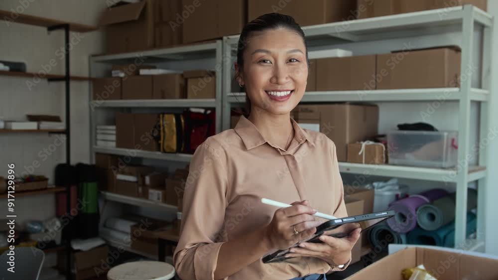 Medium portrait of cheerful female manager of Asian ethnicity filling form on tablet pc while working at storage office and smiling at camera Medium portrait of cheerful female manager of Asian ethnicity filling form on tablet pc while working at storage office and smiling at camera
