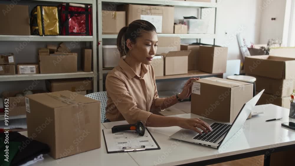 Wide shot of female Asian manager scanning parcels in boxes while working on laptop at desk in storage office Wide shot of female Asian manager scanning parcels in boxes while working on laptop at desk in storage office