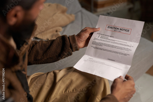 Person holding eviction notice while sitting on couch and reading in a dimly lit room without natural light coming in through the window behind photo