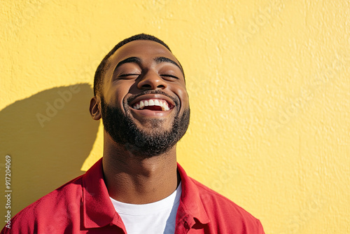 Black Man Expressing Joyful Laughter in a Close-Up Headshot, Sunlight Highlighting His Face, Red Shirt and White T-Shirt Against a Yellow Wall