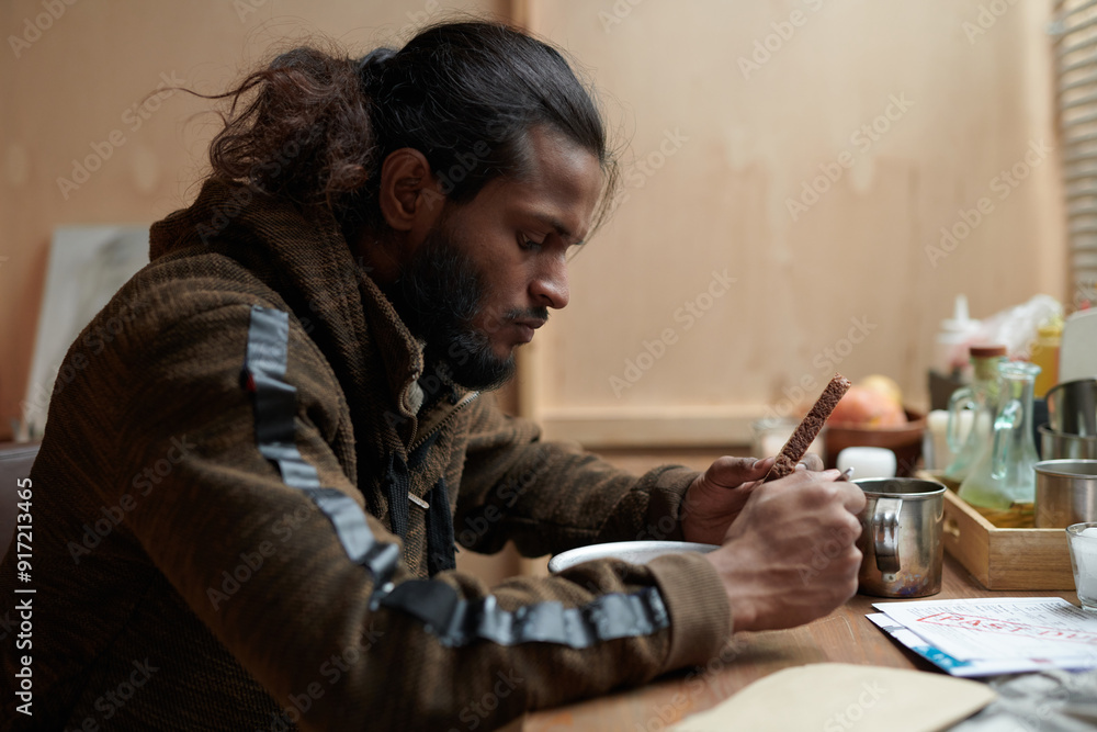 Man deeply engrossed in his work while sitting at table in warmly lit setting with various items placed around. Intense concentration displayed on man's face Man deeply engrossed in his work while sitting at table in warmly lit setting with various items placed around. Intense concentration displayed on man's face