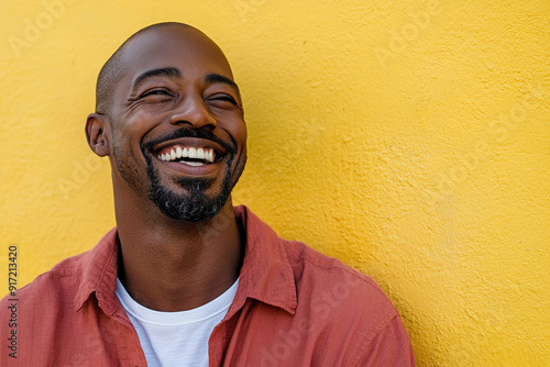 Joyful Black Man Laughing with Sunlight on His Face, Wearing a Red Shirt and White T-Shirt, Headshot Against a Bright Yellow Wall Background