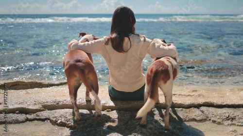 Young woman is sitting on the rocks by the sea with her two dogs, embracing them, enjoying a moment of peace and tranquility