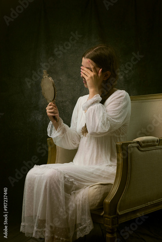 young woman in white vintage dress sitting in an armchair, holding an antique mirror and covering her face with her other hand III photo