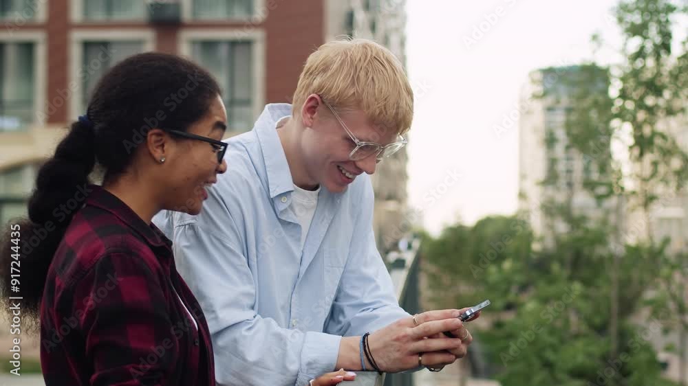 Young caucasian guy and a young black girl looking at a smartphone, becoming extremely overjoyed and laughing outdoors with a building as a backdrop Young caucasian guy and a young black girl looking at a smartphone, becoming extremely overjoyed and laughing outdoors with a building as a backdrop