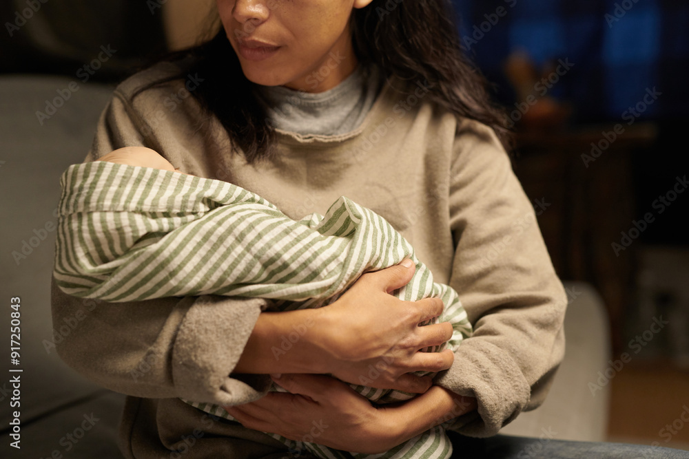 Fototapeta premium A mother gently cradling her newborn baby wrapped in striped blanket while sitting on couch, with soft lighting highlighting a serene scene