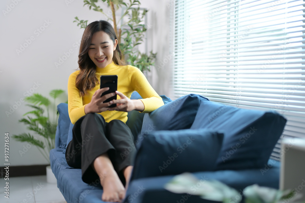 Young Woman Relaxing on a Blue Sofa Using Smartphone in a Bright Modern Living Room with Indoor Plants Young Woman Relaxing on a Blue Sofa Using Smartphone in a Bright Modern Living Room with Indoor Plants