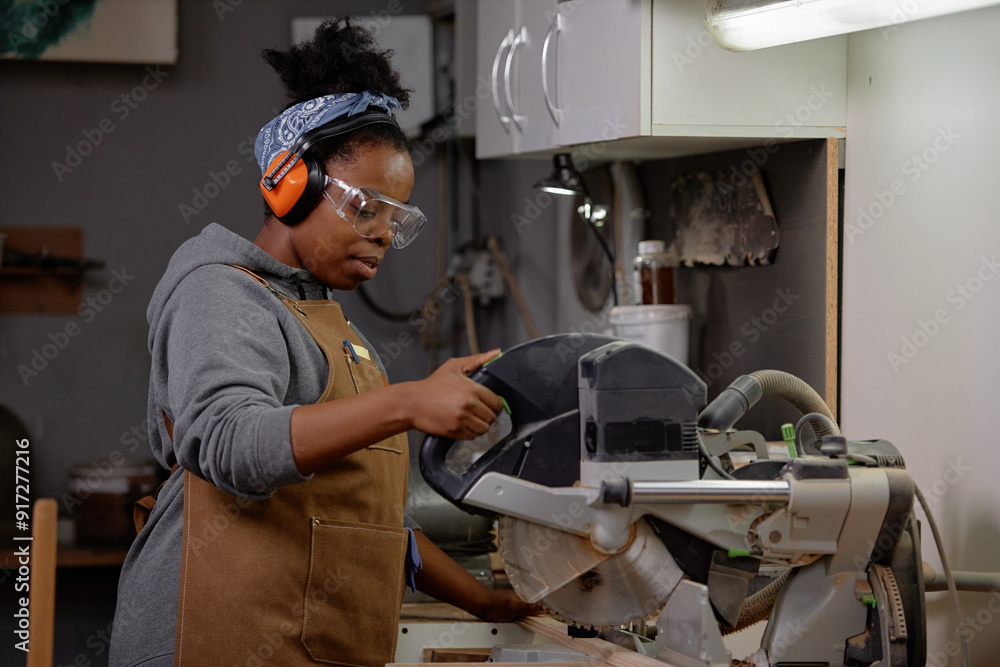 Woman wearing safety goggles and earmuffs operating circular saw in workshop. Person wearing work apron indicative of hands-on practical environment Woman wearing safety goggles and earmuffs operating circular saw in workshop. Person wearing work apron indicative of hands-on practical environment