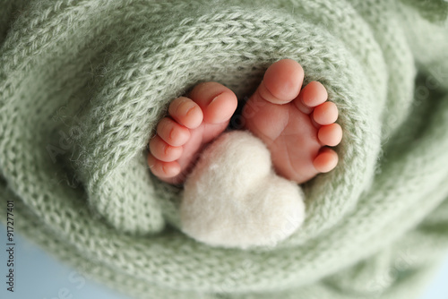The tiny foot of a newborn baby. Soft feet of a new born in a olive green wool blanket. Close up of toes, heels and feet of a newborn. Knitted white heart in the legs of a baby. Macro photography. 