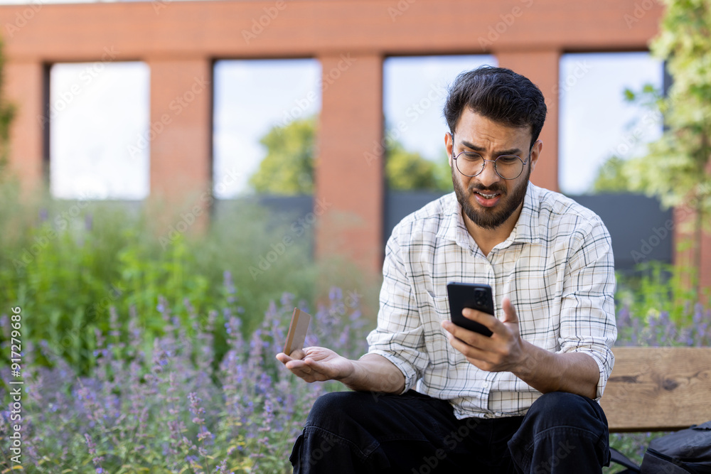 Obraz premium Worried and frustrated young indian man wearing glasses sitting outside on a bench and looking upset at the mobile phone screen while waving his hand incoherently