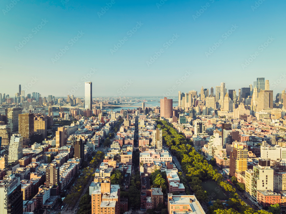 Fototapeta premium Sunrise above Lower East Manhattan buildings, park and street leading to Manhattan Bridge. Clear sky, morning light. Brooklyn Bridge on the horizon