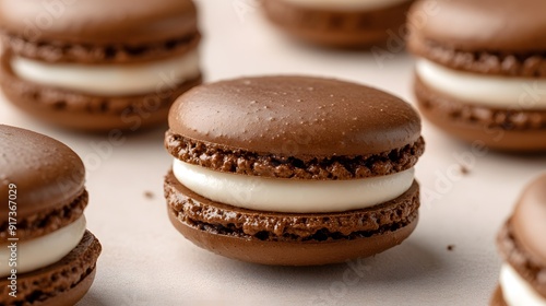 A close-up of chocolate macarons with white cream filling, placed on a light beige surface. The macarons have glossy and slightly smooth surfaces, showcasing their delicate texture