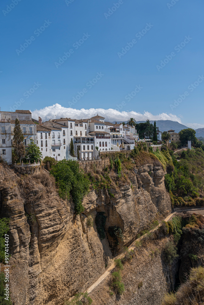Naklejka premium A stunning view of whitewashed houses perched on the edge of the Tajo Gorge in Ronda, Spain, with rugged cliffs and a clear blue sky. Perfect for themes of travel, architecture, and scenic landscapes.