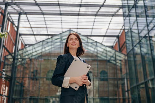 A confident businesswoman is standing in modern glass architecture, holding documents
