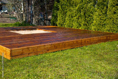 Quadro su tela Freshly stained wooden deck with a central sandpit in a backyard surrounded by t
