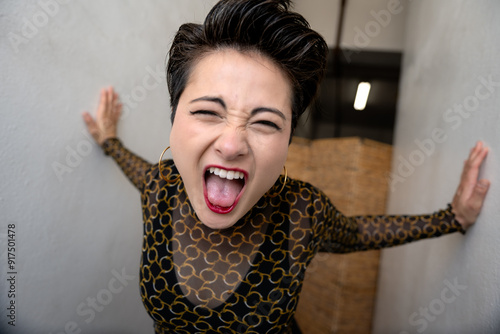 A woman with short hair and red lipstick opens her mouth wide in excitement, posing in a narrow hallway and wearing a black sheer top with a gold chain pattern. photo