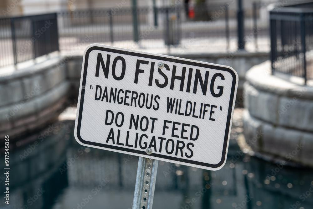 A black and white metal sign with no fishing, dangerous wildlife, do not feed alligators. The signage is on a metal post with water in the background. The signboard warns anglers of wild animals. A black and white metal sign with no fishing, dangerous wildlife, do not feed alligators. The signage is on a metal post with water in the background. The signboard warns anglers of wild animals.