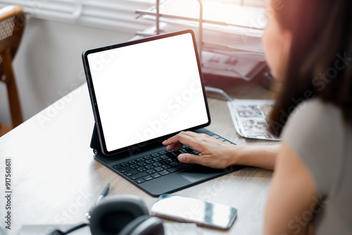 A woman uses a tablet with a keyboard at a home office desk, surrounded by various items, in bright sunlight.