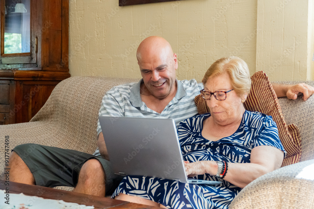 One man with his retired mother working together at home using a laptop. One man with his retired mother working together at home using a laptop.