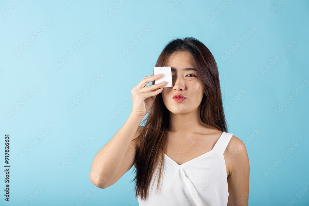 Fototapeta premium Facial Beauty Treatment Hygiene skin. Asian beautiful young woman removing cosmetic make up using pad cotton face cleaner disc, studio shot isolated on blue background, Happy female cleaning eyes