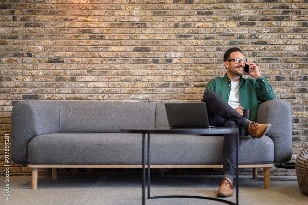 Happy male programmer with coffee cup discussing ideas over phone call while sitting on sofa in office Happy male programmer with coffee cup discussing ideas over phone call while sitting on sofa in office