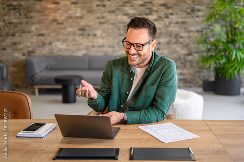 Male computer programmer smiling and discussing project details over video call on laptop in office photo