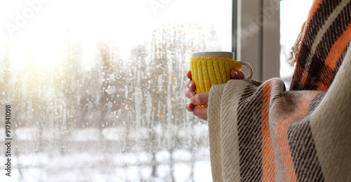 unrecognizable man in a blanket with a mug in his hands against the background of a window with raindrops on a cold day. warming atmosphere with a hot drink