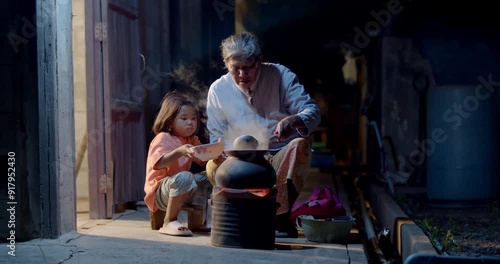 A 5-year-old Asian girl, the daughter of a poor single mother in rural Thailand, waits for food as her grandmother cooks. The scene highlights the simplicity and struggles of life in Southeast Asia.
