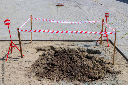 The construction site is decorated with wooden barriers with red and white tape covering the newly dug hole. Safety signs indicate restricted access, ensuring public awareness of nearby construction a