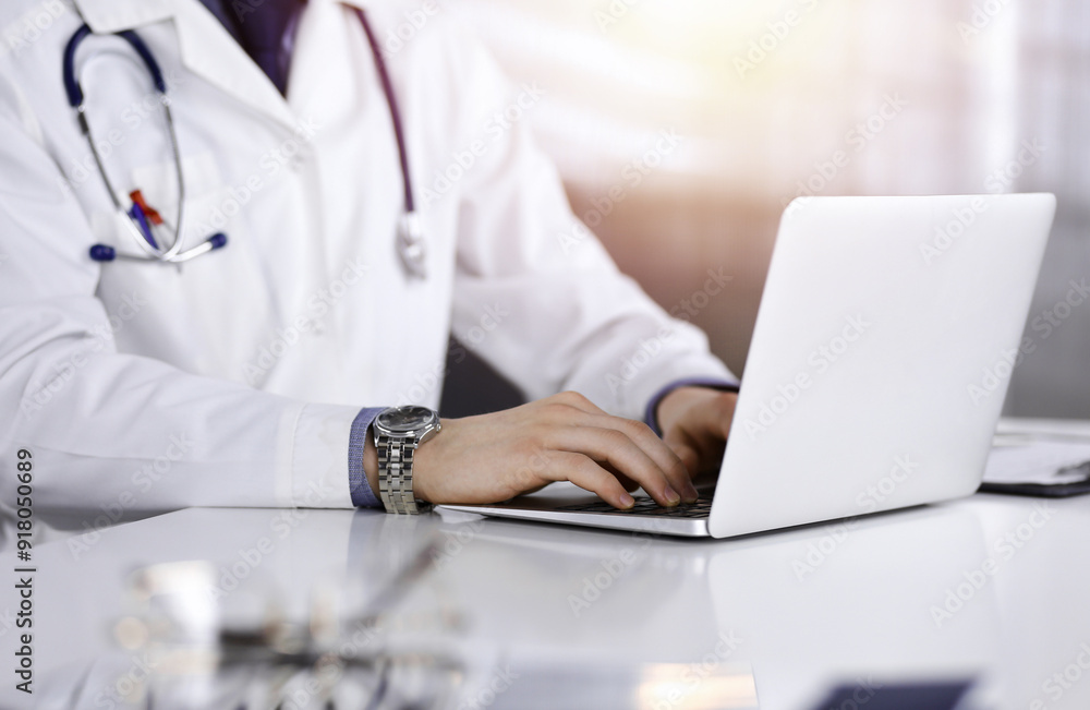 Unknown male doctor sitting and working with laptop in a darkened clinic, glare of light on the background, close-upof hands Unknown male doctor sitting and working with laptop in a darkened clinic, glare of light on the background, close-upof hands
