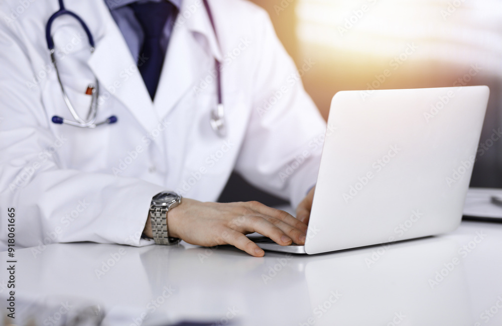 Unknown male doctor sitting and working with laptop in a darkened clinic, glare of light on the background, close-upof hands Unknown male doctor sitting and working with laptop in a darkened clinic, glare of light on the background, close-upof hands