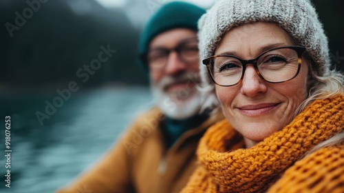 An elderly couple, warmly dressed in knit caps and sweaters, smile at the camera with a serene lake and mountains blurred in the background, symbolizing enduring love and companionship. photo