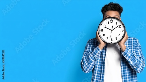 Man holding a clock in front of his face with a blue background, time management and deadline concept
 photo