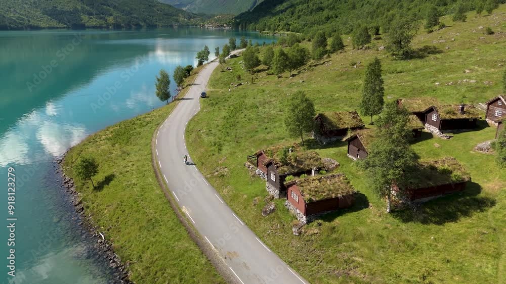 Experience a breathtaking aerial view of a winding road beside Lovatnet, surrounded by clear waters, lush greenery, and charming Norwegian huts nestled in the landscape Experience a breathtaking aerial view of a winding road beside Lovatnet, surrounded by clear waters, lush greenery, and charming Norwegian huts nestled in the landscape