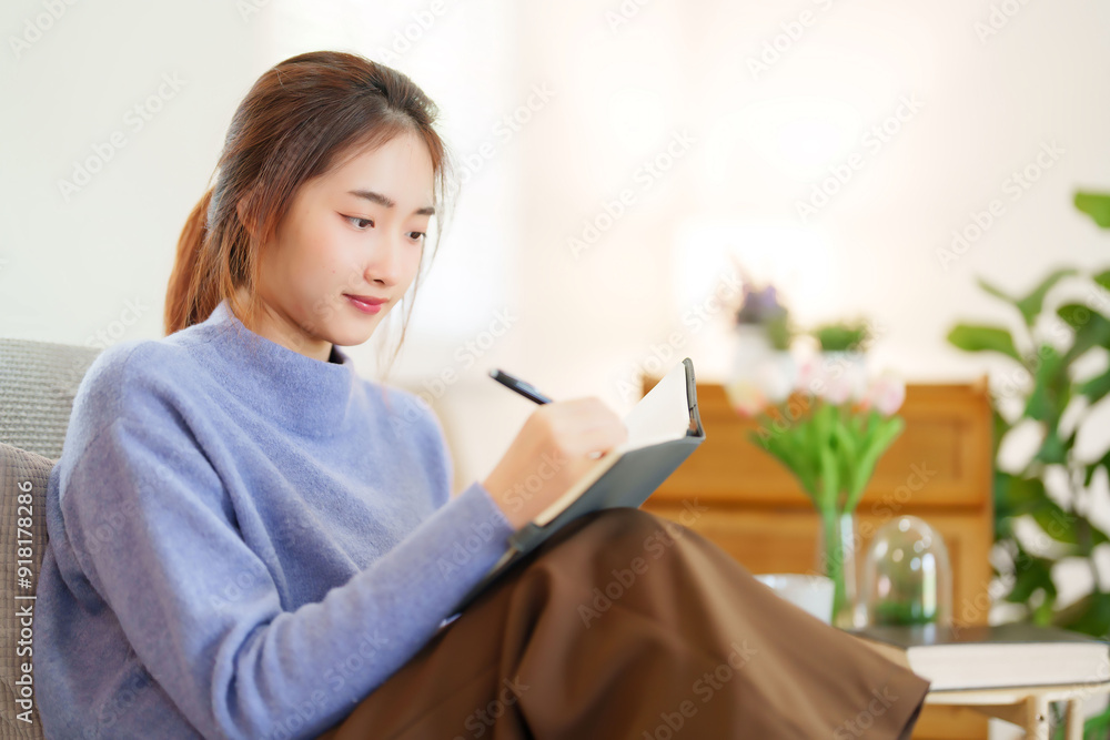 Young asian women in sweater sitting and leaning on couch to reading book and taking note for learning education while relaxation and doing activity for spending time with slow life lifestyle at home Young asian women in sweater sitting and leaning on couch to reading book and taking note for learning education while relaxation and doing activity for spending time with slow life lifestyle at home