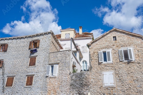 Picturesque two-story stone house with clay tiled roof, wooden shutters and balcony railing set against vivid blue sky with fluffy white clouds. Rustic, charming European architecture