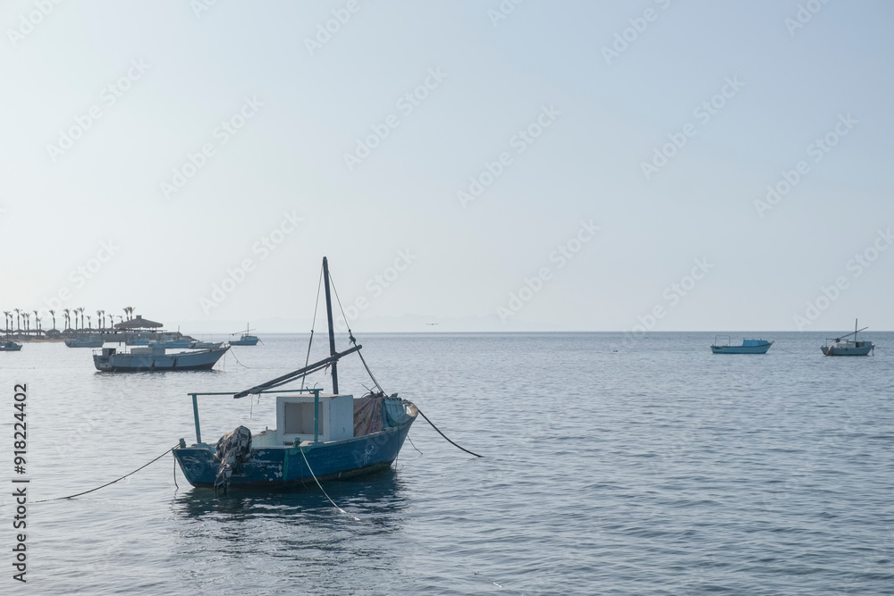 Weathered small fishing boat floats serenely on the calm sea, tethered by ropes. The distant shoreline is dotted with other boats and palm trees under a clear, bright sky Weathered small fishing boat floats serenely on the calm sea, tethered by ropes. The distant shoreline is dotted with other boats and palm trees under a clear, bright sky