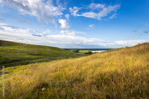 Golden grasses sway gently on rolling hills as the sun sets, casting warm light across the tranquil river nestled in the distance.