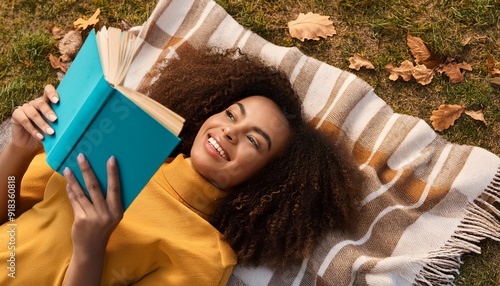 Top view of cheerful African American female with curly hair lying on blanket in park with book while smiling