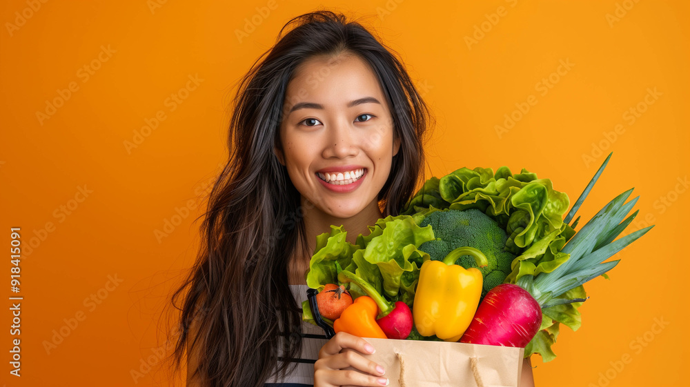 Close-up portrait of a cheerful Asian woman with long, flowing hair, holding a paper bag filled with colorful vegetables against a vibrant orange backdrop Close-up portrait of a cheerful Asian woman with long, flowing hair, holding a paper bag filled with colorful vegetables against a vibrant orange backdrop