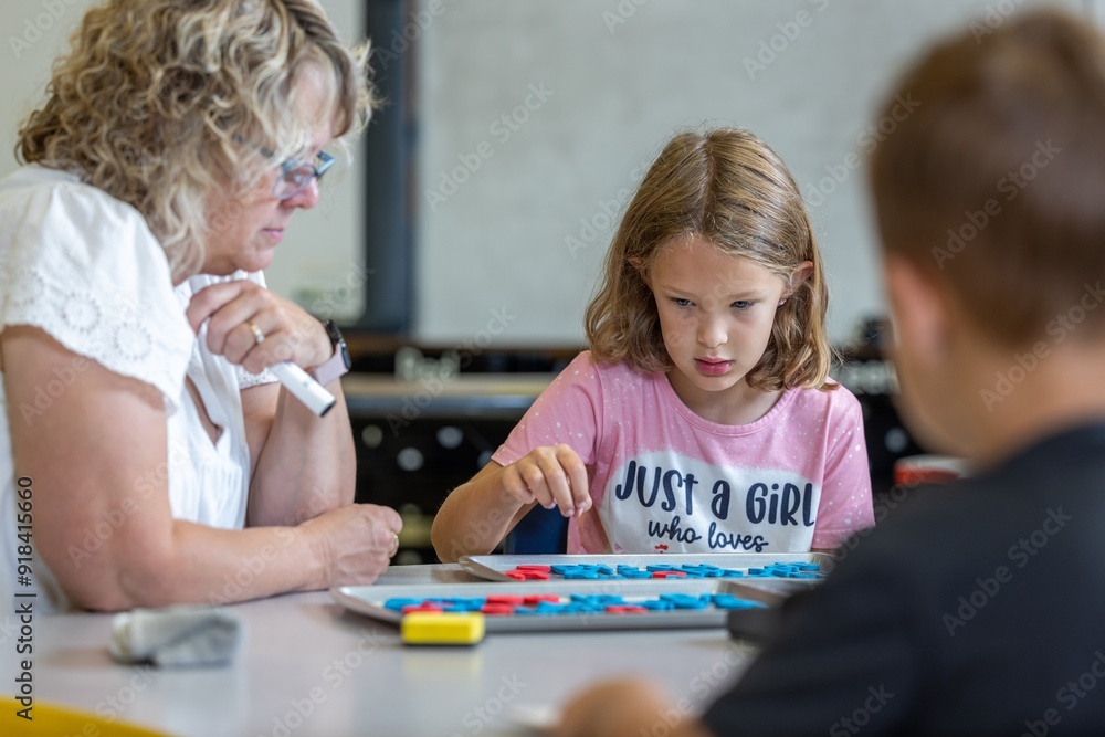 A girl in a pink shirt is sitting at a table with a woman and a boy. The girl is learning with blocks and the woman is watching her. Scene is playful and lighthearted A girl in a pink shirt is sitting at a table with a woman and a boy. The girl is learning with blocks and the woman is watching her. Scene is playful and lighthearted