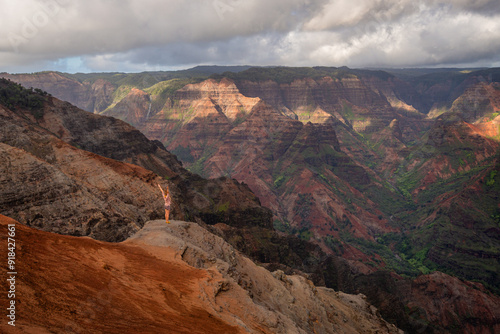 Young woman standing on the edge of cliff with hands up in air in Waimea Canyon Kauai, Hawaii USA