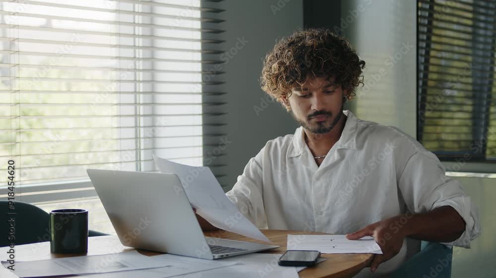 Serious young Indian businessman checking documents, analyzing financial papers, preparing audit report at workplace. Serious young Indian businessman checking documents, analyzing financial papers, preparing audit report at workplace.