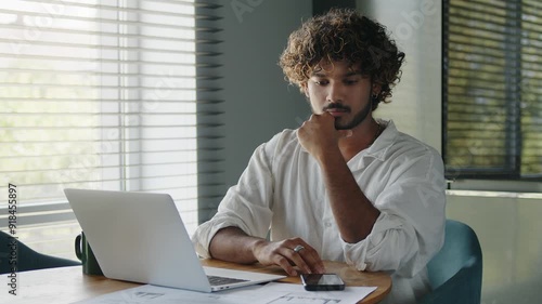 Young serious business man sits at table in home office, works with papers. 