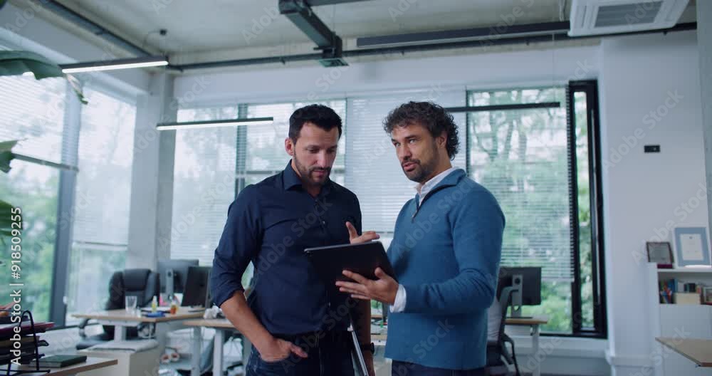 Attractive Caucasian manager holding tablet device and communicating with worker in middle of office. Busy men discussing about new business project or paths of improvement. Partnership concept. Attractive Caucasian manager holding tablet device and communicating with worker in middle of office. Busy men discussing about new business project or paths of improvement. Partnership concept.