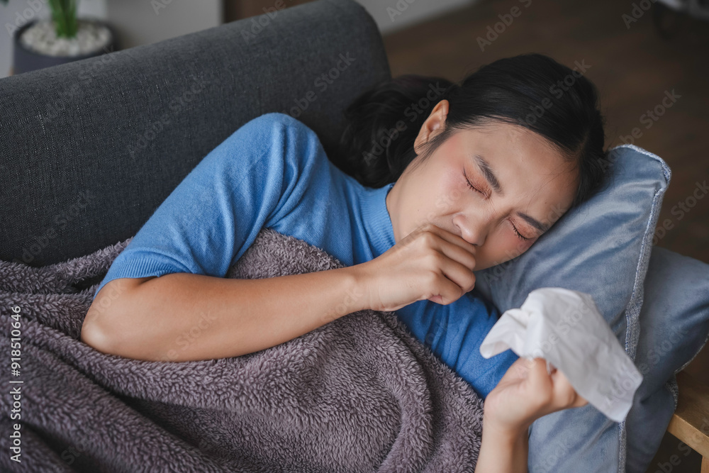 Young woman is suffering from a cold, lying on the sofa, coughing, and blowing her nose with a tissue Young woman is suffering from a cold, lying on the sofa, coughing, and blowing her nose with a tissue