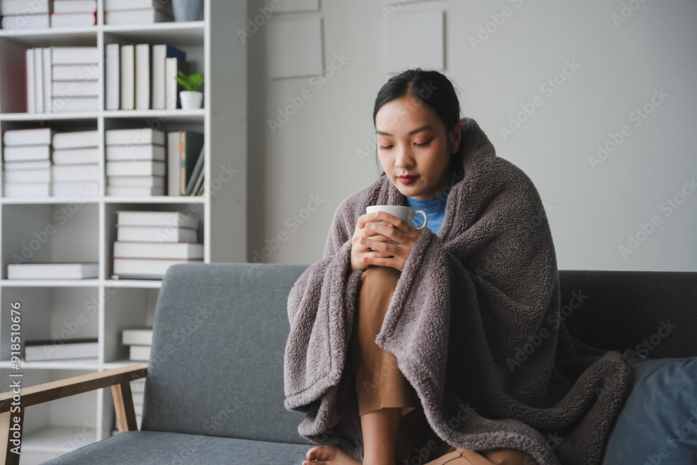 Young woman is sitting on a sofa wrapped in a blanket, holding a mug and feeling unwell Young woman is sitting on a sofa wrapped in a blanket, holding a mug and feeling unwell