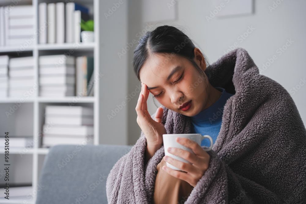 Young asian woman feeling sick having headache holding a mug with hot drink sitting on a sofa at home Young asian woman feeling sick having headache holding a mug with hot drink sitting on a sofa at home