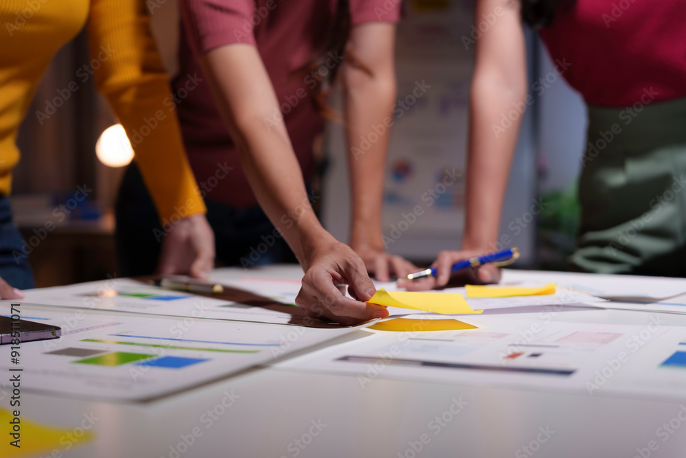 Fototapeta premium Team is working late at night, using colorful sticky notes to organize their work on a big desk full of graphs and analysis