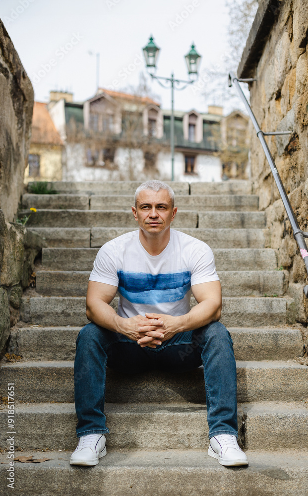 A man in a white shirt and blue jeans sits on a set of stairs. He is looking at the camera with a serious expression. Concept of solitude and contemplation A man in a white shirt and blue jeans sits on a set of stairs. He is looking at the camera with a serious expression. Concept of solitude and contemplation